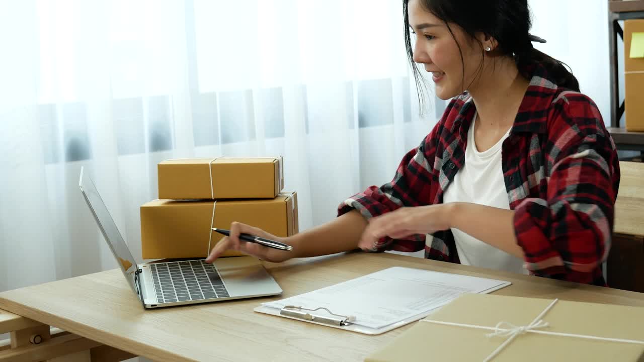 网上卖家近距离接触年轻的亚洲女子，她正在输入笔记本电脑键盘，检查网上订单，检查货物库存，发货包裹，发货邮政。亚洲女性在家办公室做小生意视频素材