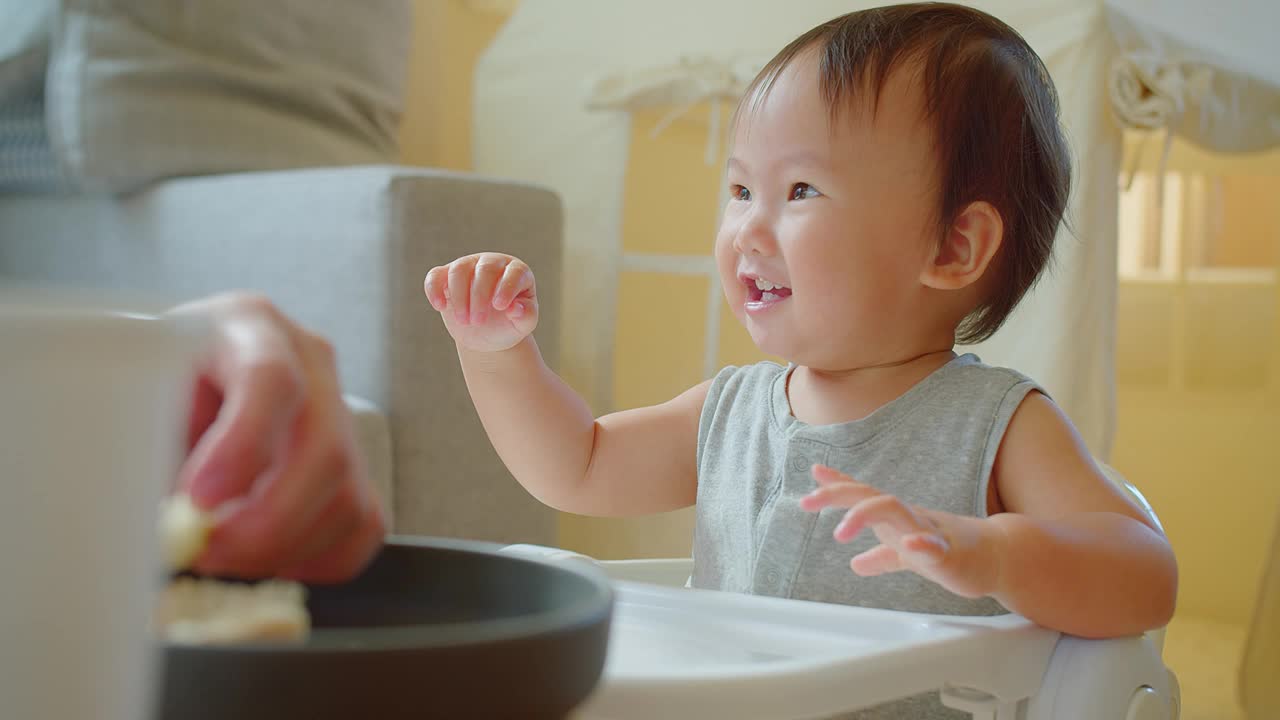 在一个阳光明媚的家庭里，一个快乐的幼儿坐在高脚椅上享受用餐时间，带着明亮迷人的微笑伸手去拿食物视频素材