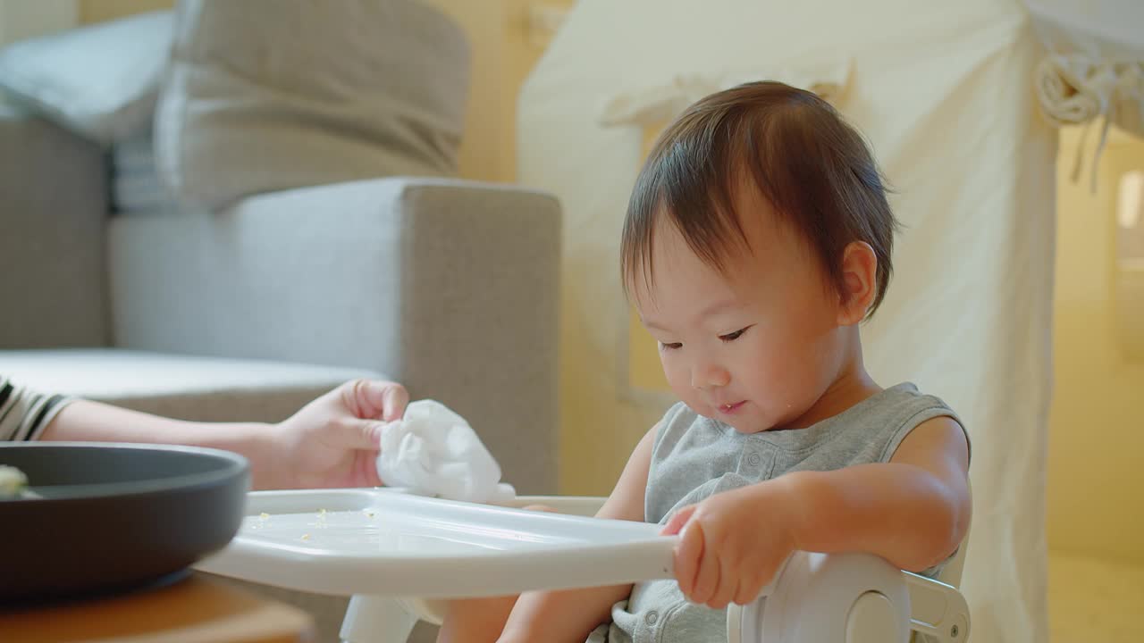 在一个阳光明媚的家庭里，一个快乐的幼儿坐在高脚椅上享受用餐时间，带着明亮迷人的微笑伸手去拿食物视频素材