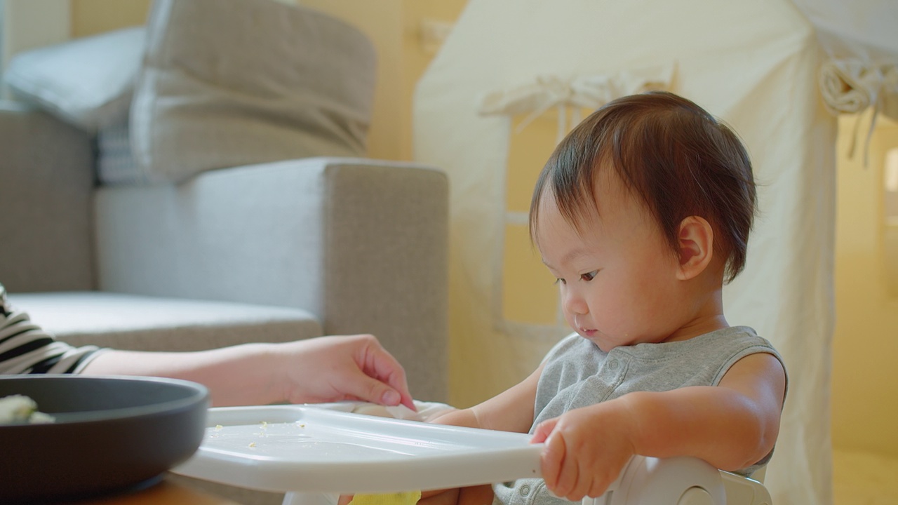 在一个阳光明媚的家庭里，一个快乐的幼儿坐在高脚椅上享受用餐时间，带着明亮迷人的微笑伸手去拿食物视频素材