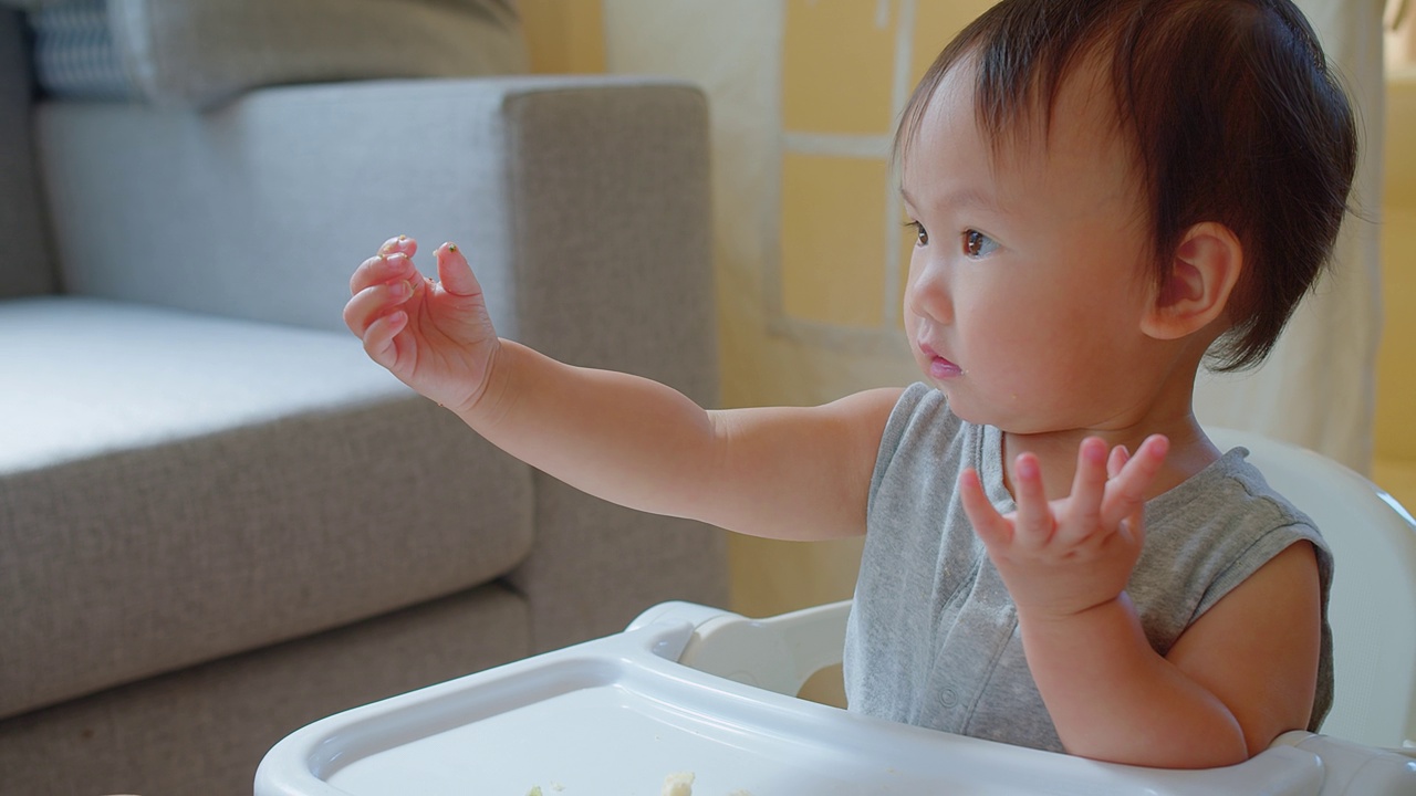 在一个阳光明媚的家庭里，一个快乐的幼儿坐在高脚椅上享受用餐时间，带着明亮迷人的微笑伸手去拿食物视频素材