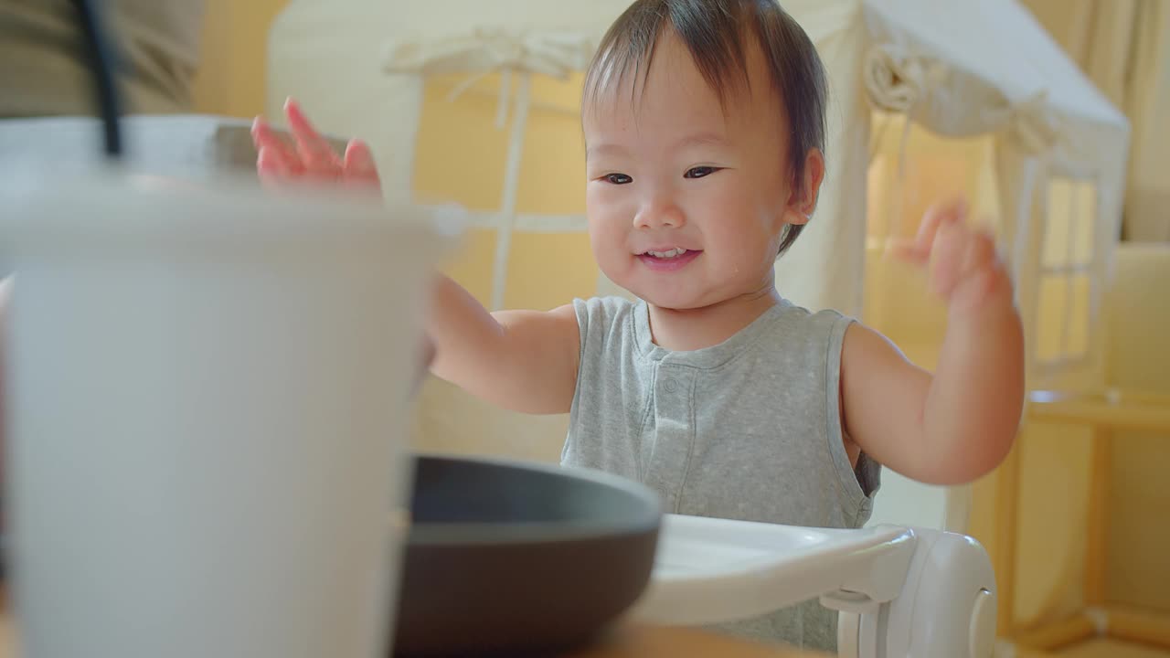 在一个阳光明媚的家庭里，一个快乐的幼儿坐在高脚椅上享受用餐时间，带着明亮迷人的微笑伸手去拿食物视频素材