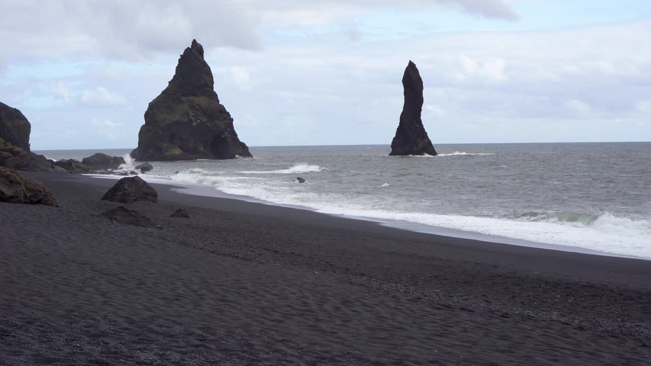 冰岛多云的天空下，黑色的沙滩上矗立着高耸的海蚀柱，海浪拍打着海岸视频素材