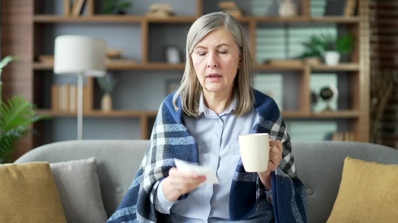 生病的老妇人裹着毯子坐在家里客厅的沙发上流鼻涕。视频素材