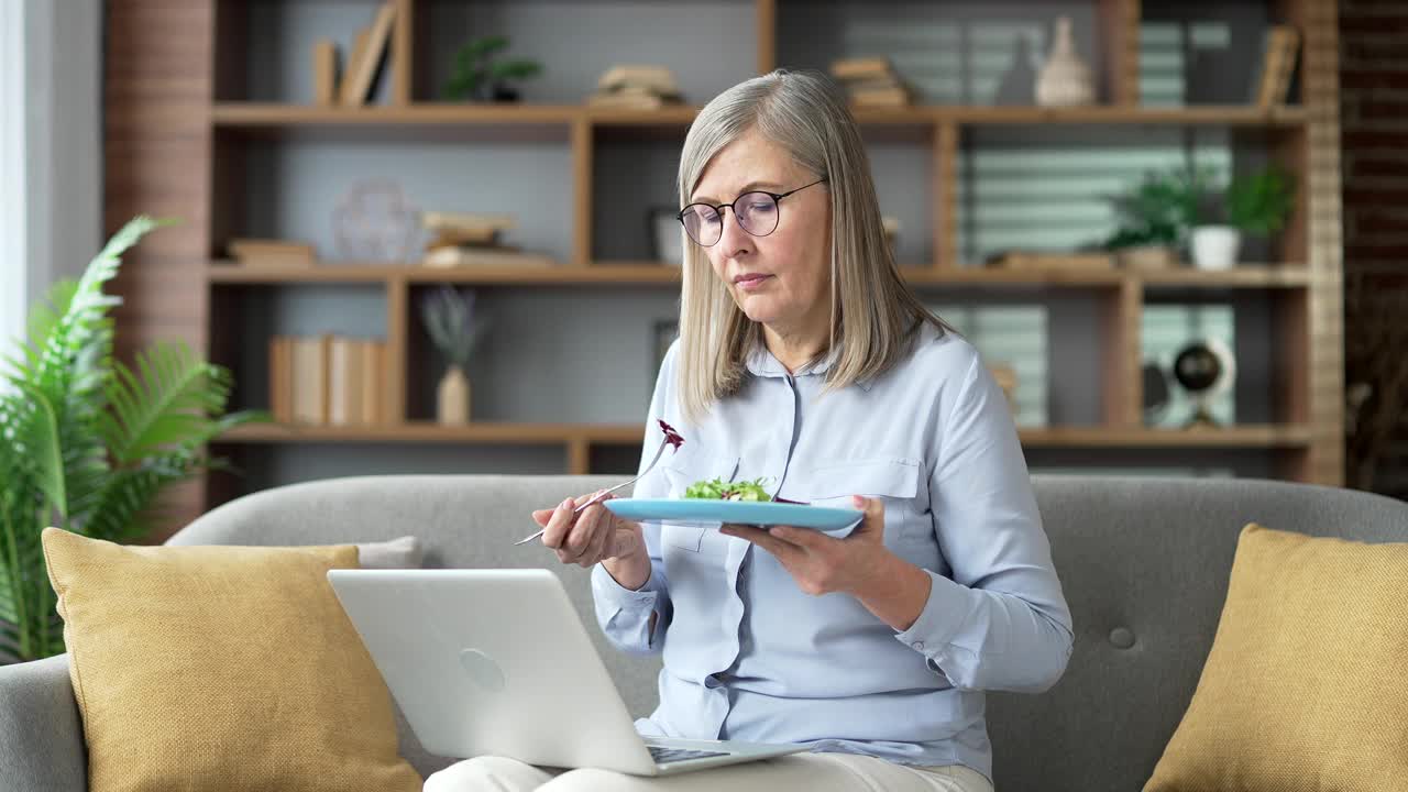 厌倦了节食减肥的老年女性带着一盘沙拉坐在家里客厅的沙发上视频素材