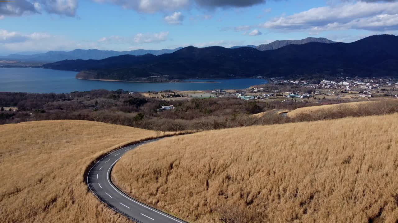 蜿蜒的道路穿过金色的田野，远处有湖景和山脉，航拍视频素材