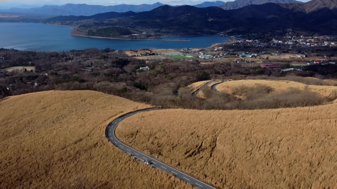 一条蜿蜒的道路穿过金色的田野，远处是湖泊和山脉，在晴朗的天空下，鸟瞰视频素材