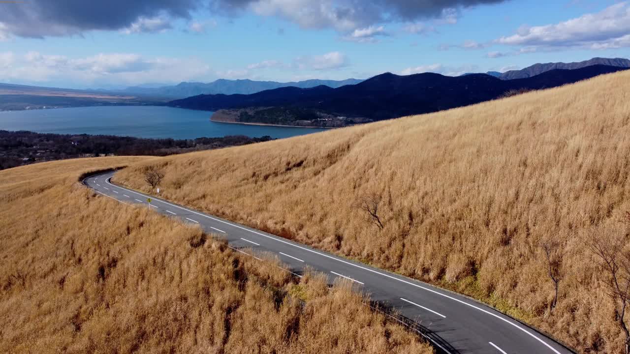 蜿蜒的道路穿过金色的山丘，可以看到湖泊和山脉，在多云的天空下，航拍视频素材