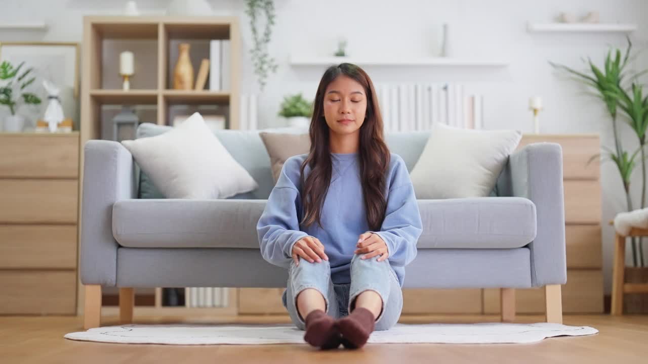 快乐的年轻亚洲女性在家里练习瑜伽和冥想，坐在客厅的地板上，以莲花的姿势，闭上眼睛放松。正念冥想和幸福概念视频素材