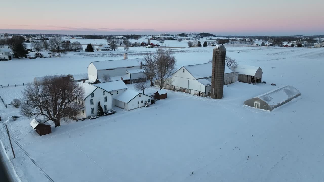 黄昏时鸟瞰图，一个白雪覆盖的农场，有多座建筑物和一个筒仓。降雪后的无人机轨道。美国乡村的日出。视频素材