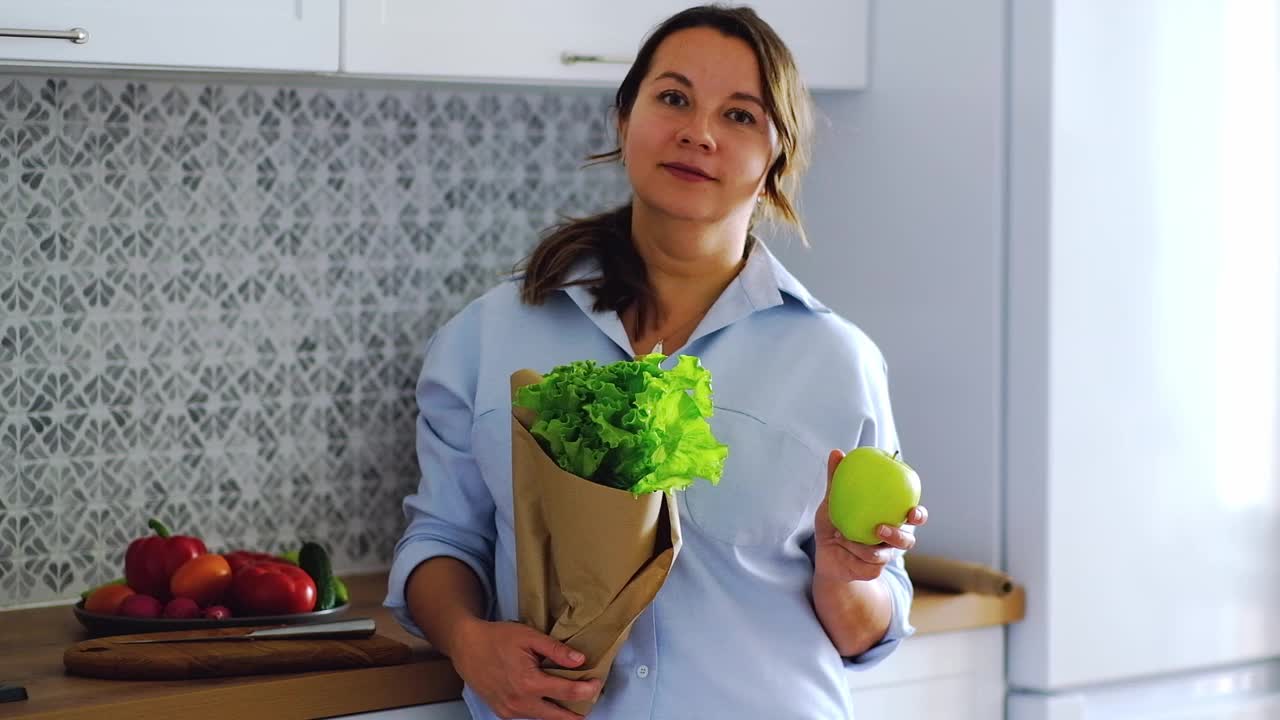 一个年轻漂亮的女人手里拿着新鲜的生菜和苹果。低碳水化合物饮食和健康饮食。在厨房烹饪美味的素食视频素材