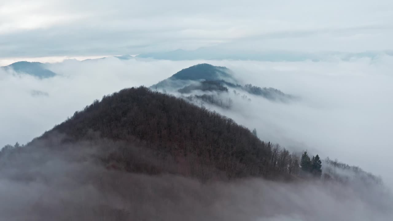 云雾缭绕的山峦和被森林覆盖的山坡透过厚厚的云层隐约可见，这是黎明时分的航拍照片视频素材