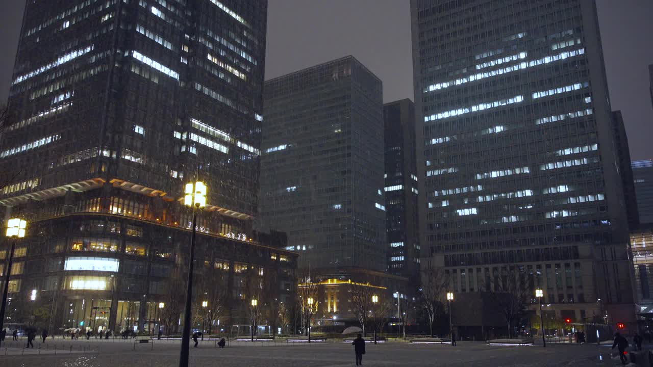 夜晚，东京的摩天大楼上飘着雪花，丸之内的行人打着伞行走视频下载