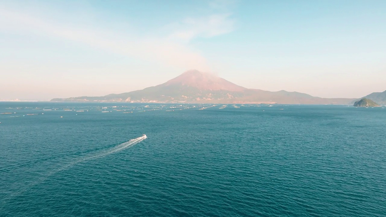 在阳光明媚的日子里，一艘渔船驶向鹿儿岛湾蔚蓝的海水和冒着烟的樱岛火山视频素材