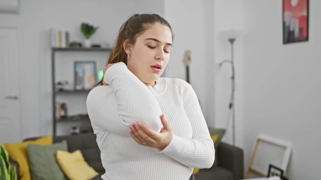 穿着白色毛衣的年轻西班牙妇女在舒适的室内经历肘部疼痛视频素材