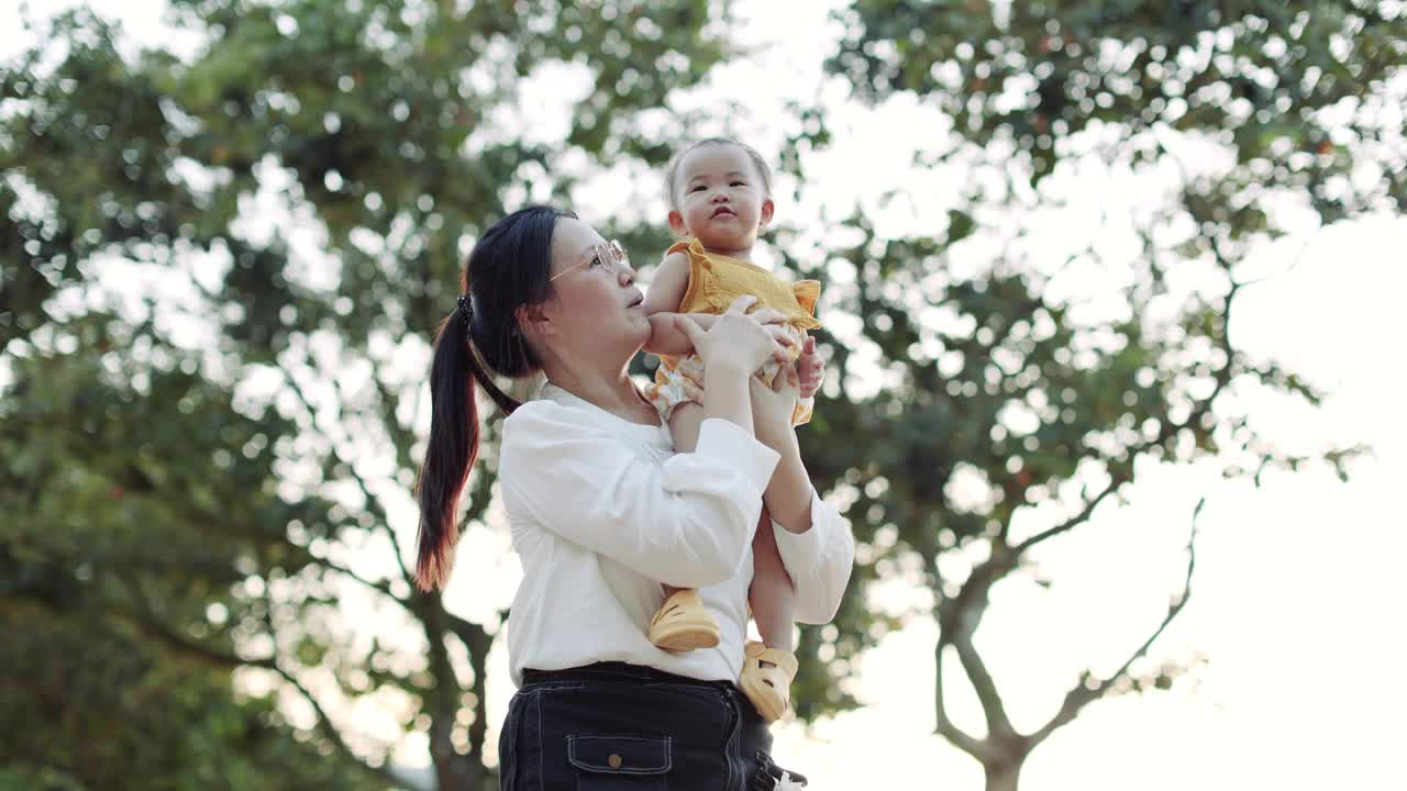 一位亚洲母亲把女儿扛在肩上，在花园公园里给她挠痒痒。库存图片视频素材