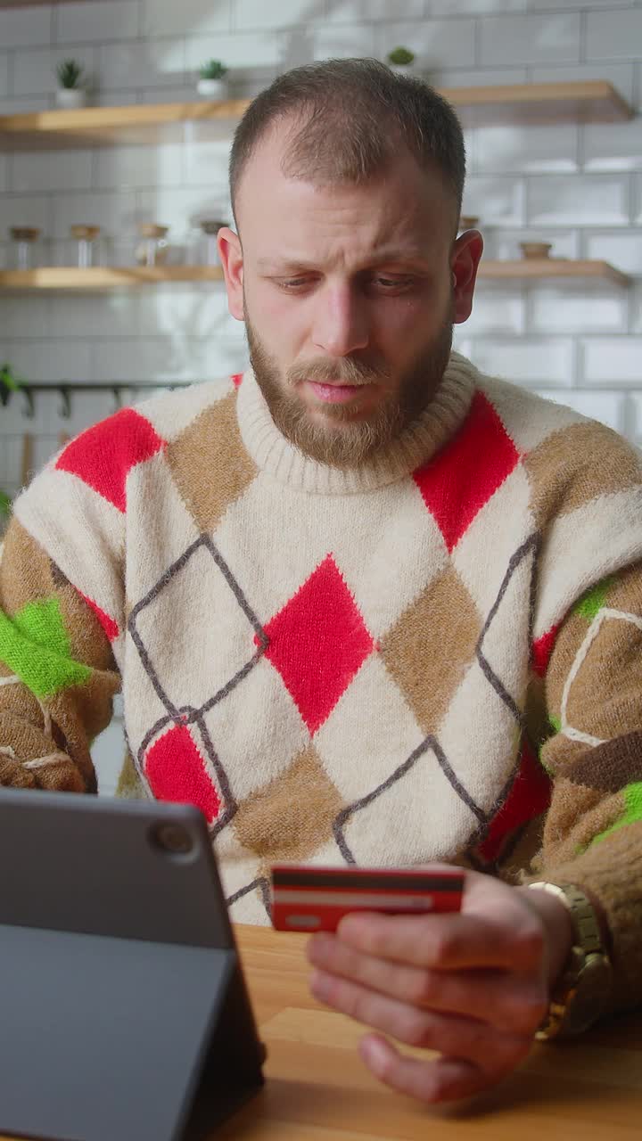 垂直视频。微笑的男人在平板电脑上输入信用卡号码进行网上购物，坐在家里的厨房里就可以安全方便地进行远程电子支付视频素材