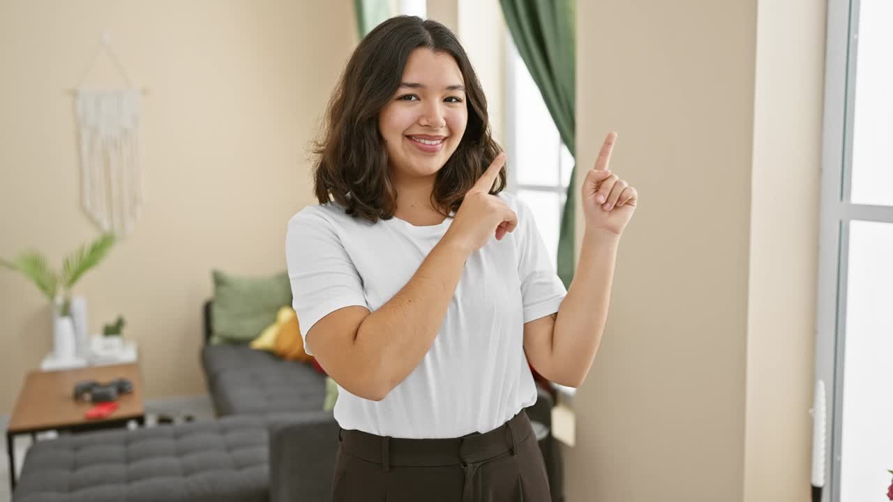 开朗的年轻西班牙女子站在家里，自信地指着一边微笑。准备好迎接了!拍下她灿烂的笑容!视频素材