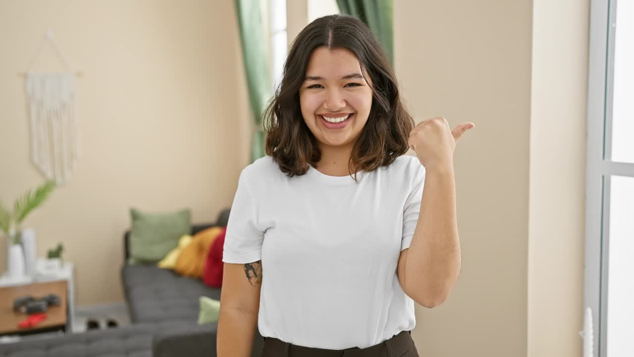 活泼的年轻西班牙裔女子站在她舒适的客厅里，张开嘴，露出灿烂的微笑，自信地用拇指指着一边。散发出纯粹的快乐、积极和信任。视频素材