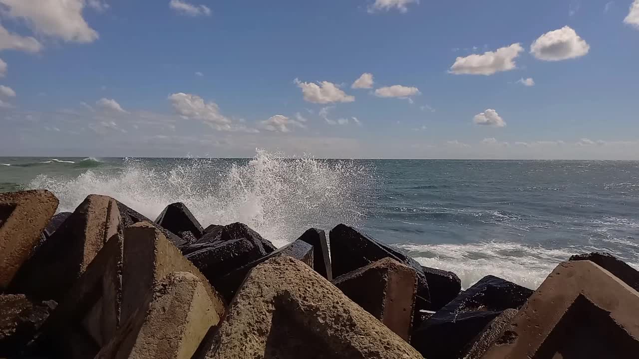 海浪冲击着岸边的石制防波堤。海浪溅在海上的码头上。海面上刮着清新的风，波涛汹涌的海面上有长长的波浪和白浪。阳光明媚的夏日户外海景。视频下载