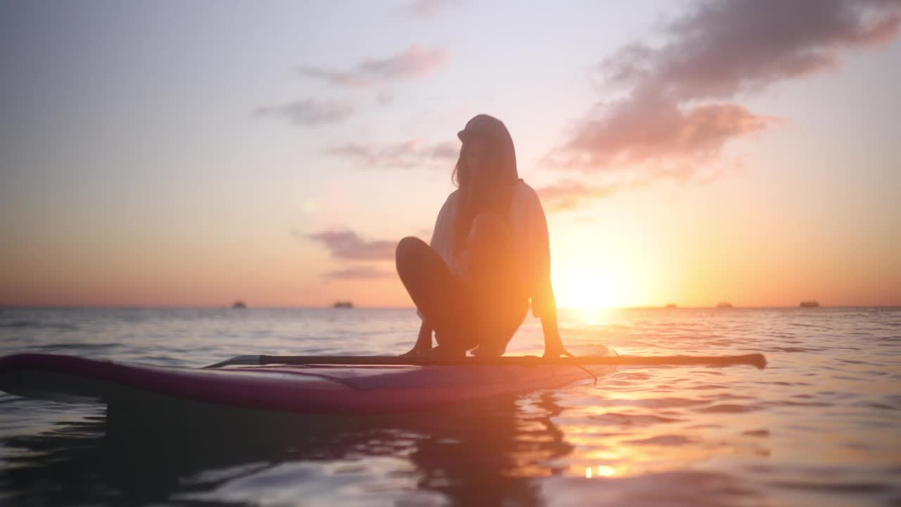 一名女子坐在平静的海面上的站立式桨板上欣赏日落视频素材