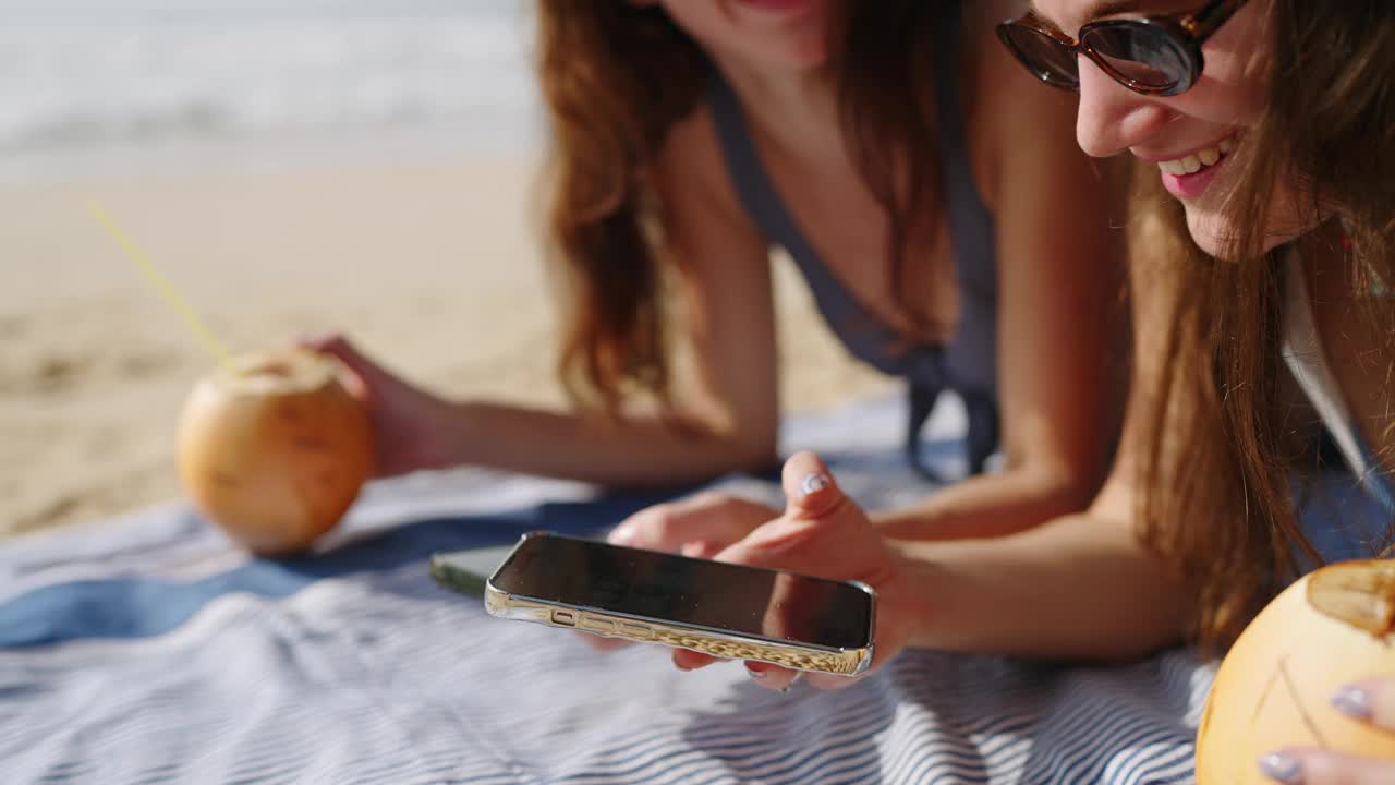 快乐的女孩晒黑，在沙滩上用手机上网。快乐的女士们在海边晒日光浴，用手机发短信。女人喝椰子汁。暑假对社交媒体上瘾。特写镜头。视频素材