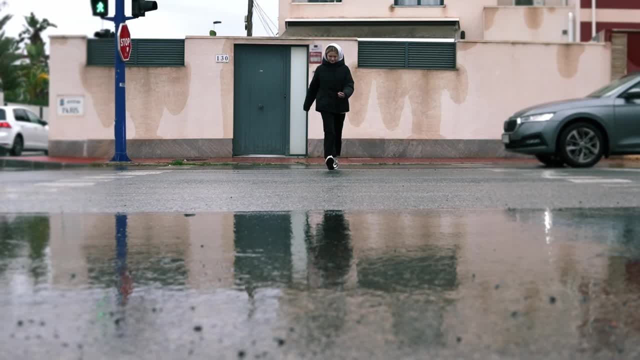 白天，一个十几岁的女孩穿着黑白运动鞋，冒雨在红绿灯前穿过街道上人行道上的一个大水坑，长长的特写。溅起的水花落在受影响的各方身上。一边的计划。视频素材