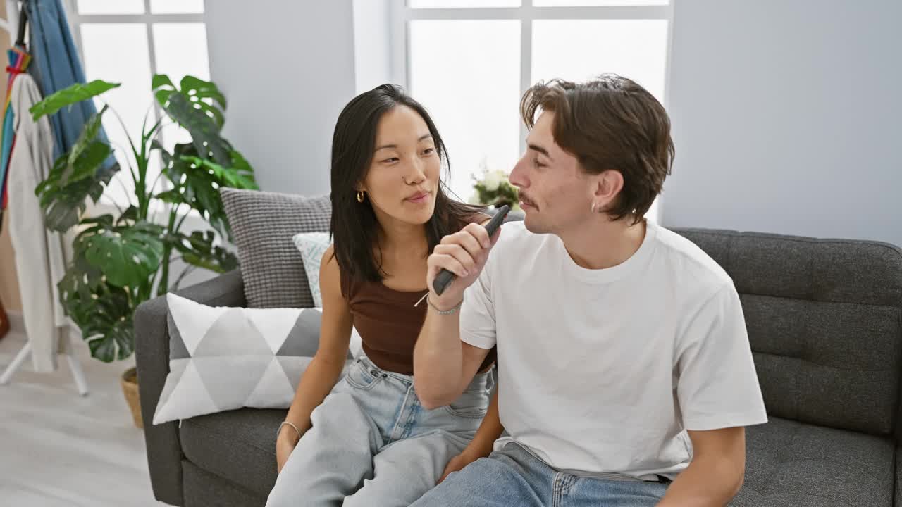 不同种族的夫妇在家里唱歌，亚洲女人用电视遥控器当麦克风，而男人微笑着。视频素材