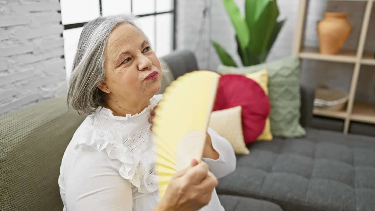 一个成熟的女人在舒适、装饰精美的客厅里拿着一把黄色的扇子降温，散发着优雅和舒适的气息。视频素材