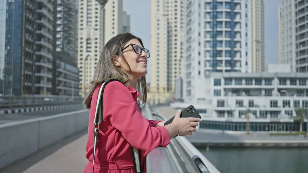 微笑的女人戴着眼镜，穿着粉色衬衫，拿着电话，背景是迪拜码头的摩天大楼视频素材