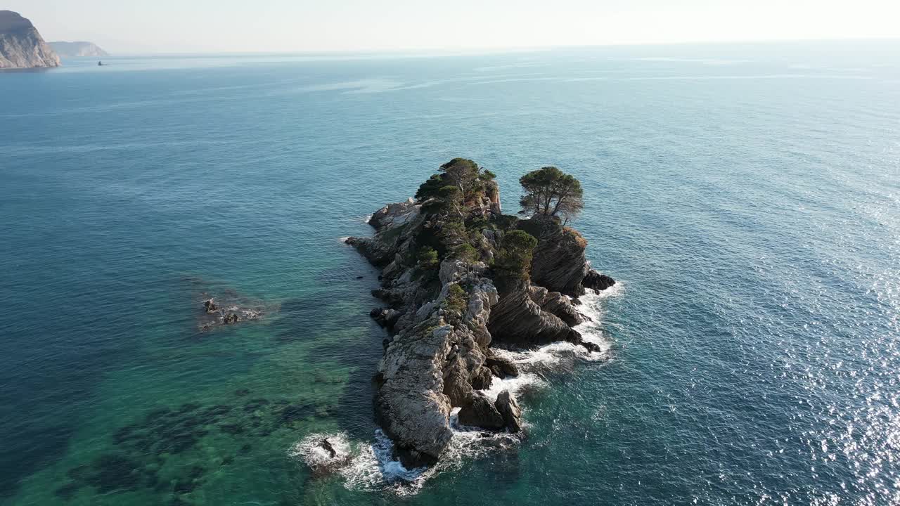 在一个阳光明媚的日子里，一架无人机在海中央一个有树的岩石岛上空飞行视频素材