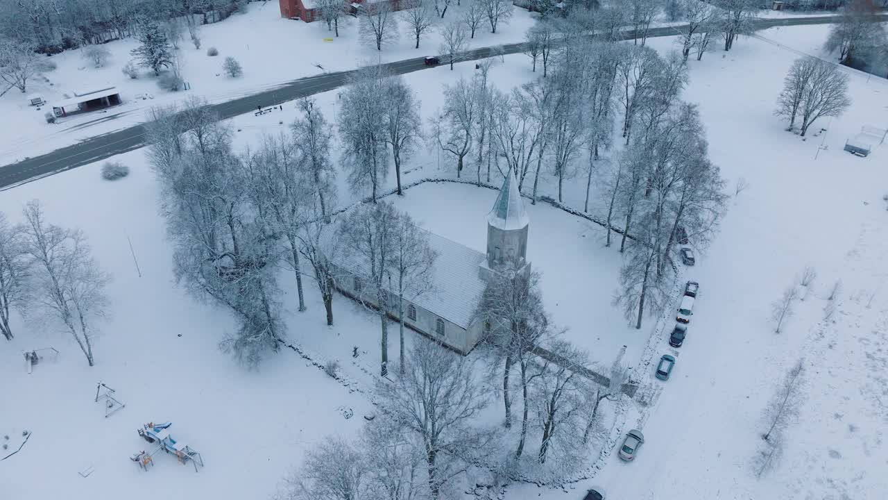 在Renda村(Kuldiga教区，拉脱维亚)被雪覆盖的教堂周围光秃秃的树木，鸟瞰，向前移动，倾斜向下视频素材