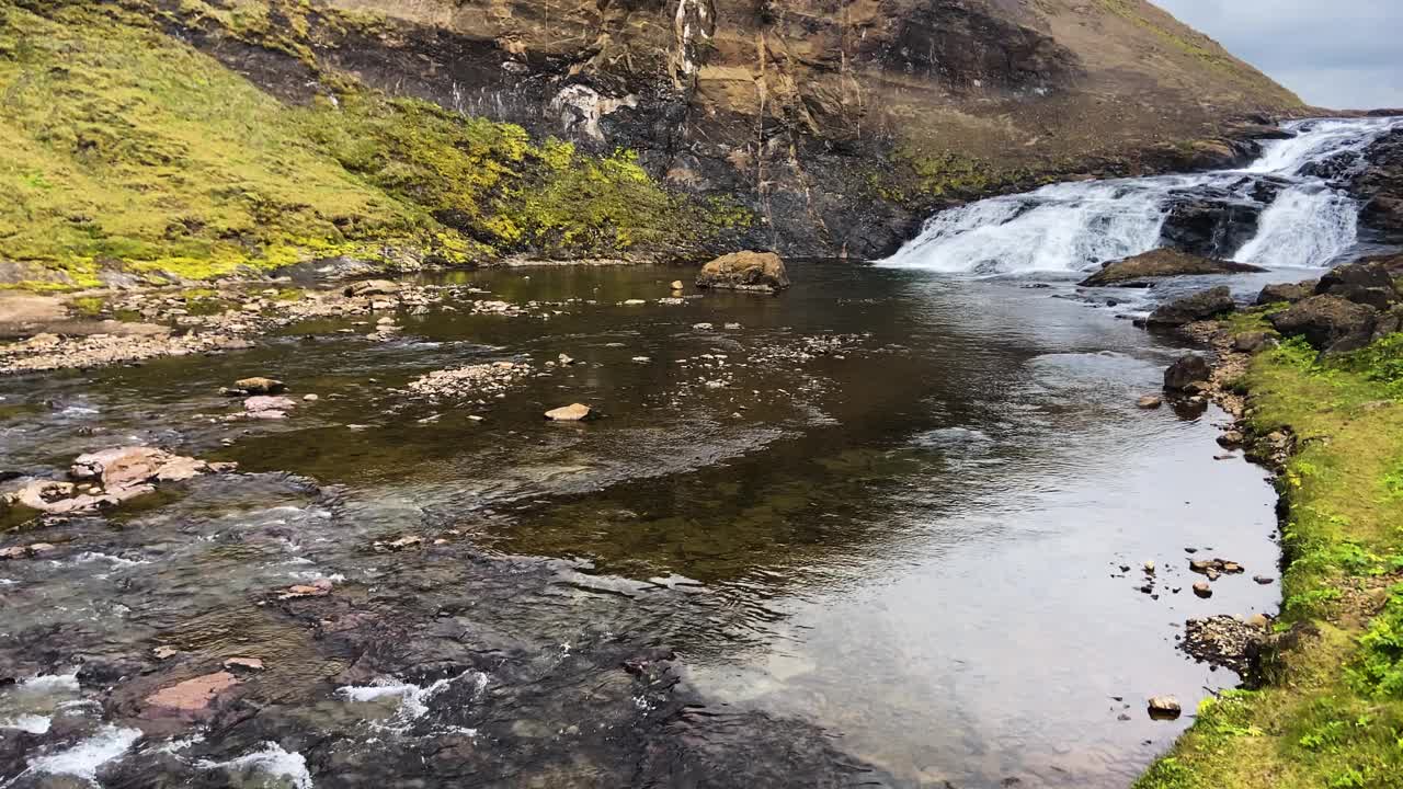 平缓的河流在冰岛第二高的瀑布格列默瀑布前流淌，周围环绕着绿色的苔藓悬崖视频素材