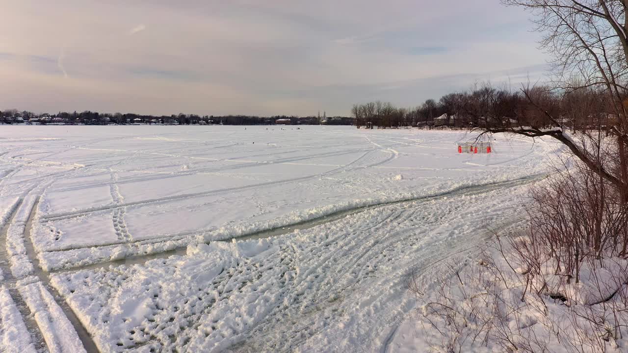 在拉瓦尔西部，从低角度俯瞰结冰的河上的白色积雪，可以看到轮胎的痕迹，并向一群在冰上玩耍的人移动视频素材