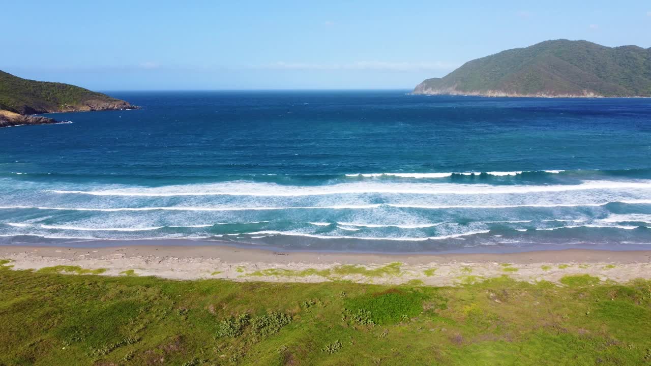 一个充满活力的海滩，海浪拍打，周围是绿色植物和山丘，在晴朗的天空下，鸟瞰图视频素材