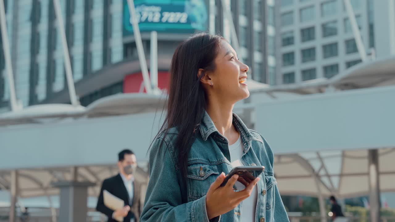 年轻的亚洲女子穿着牛仔裤，夹克，衣服，在晴朗的日子里走在城市的街道上，用智能手机发短信视频素材