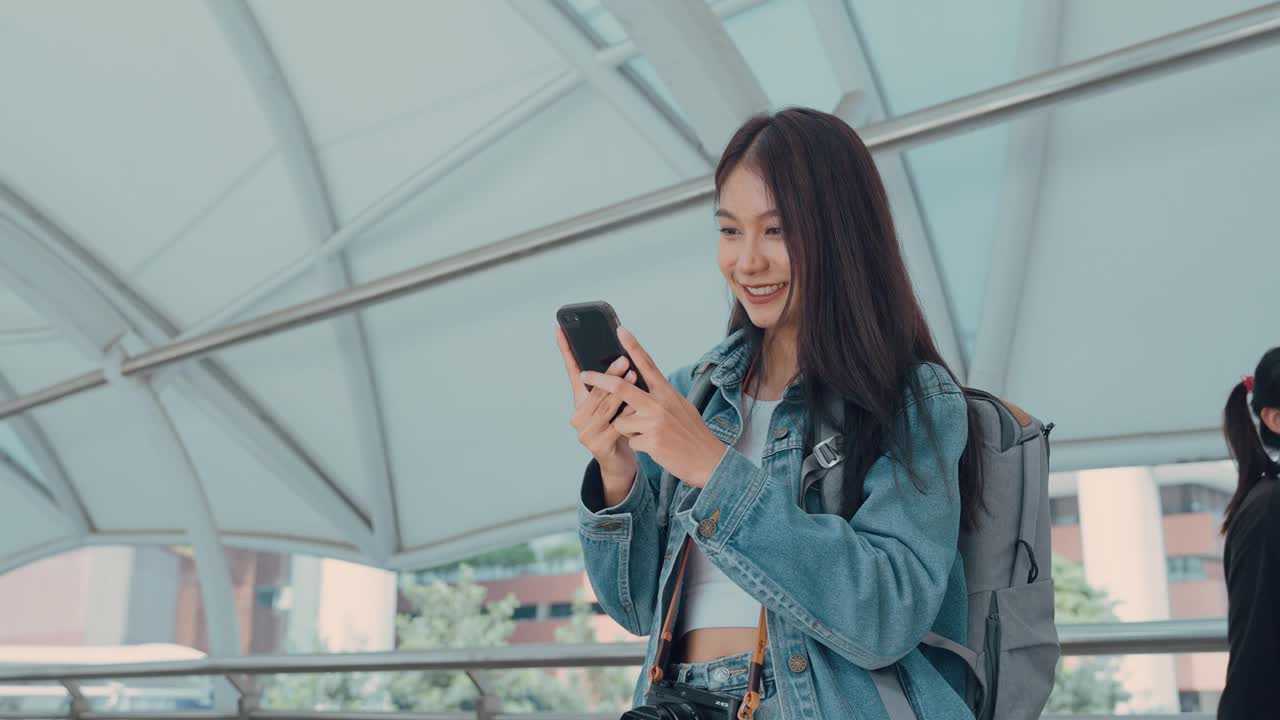 年轻的亚洲女子微笑着，在户外使用移动智能手机。穿着牛仔裤夹克拿着智能手机出现在公共城市的快乐美女。在线技术与传播理念视频素材