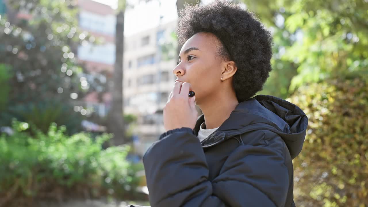 一位体贴的卷发非裔美国妇女戴着无线耳机站在绿树成荫的公园里。视频素材