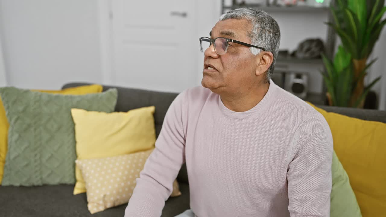 一个成熟的男人坐在家里的沙发上，一副若有所思的表情，周围是五颜六色的靠垫和室内植物。视频素材