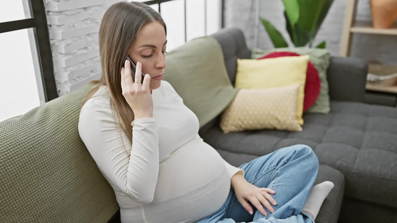 身着便服的西班牙孕妇坐在家里的沙发上讲手机。视频素材