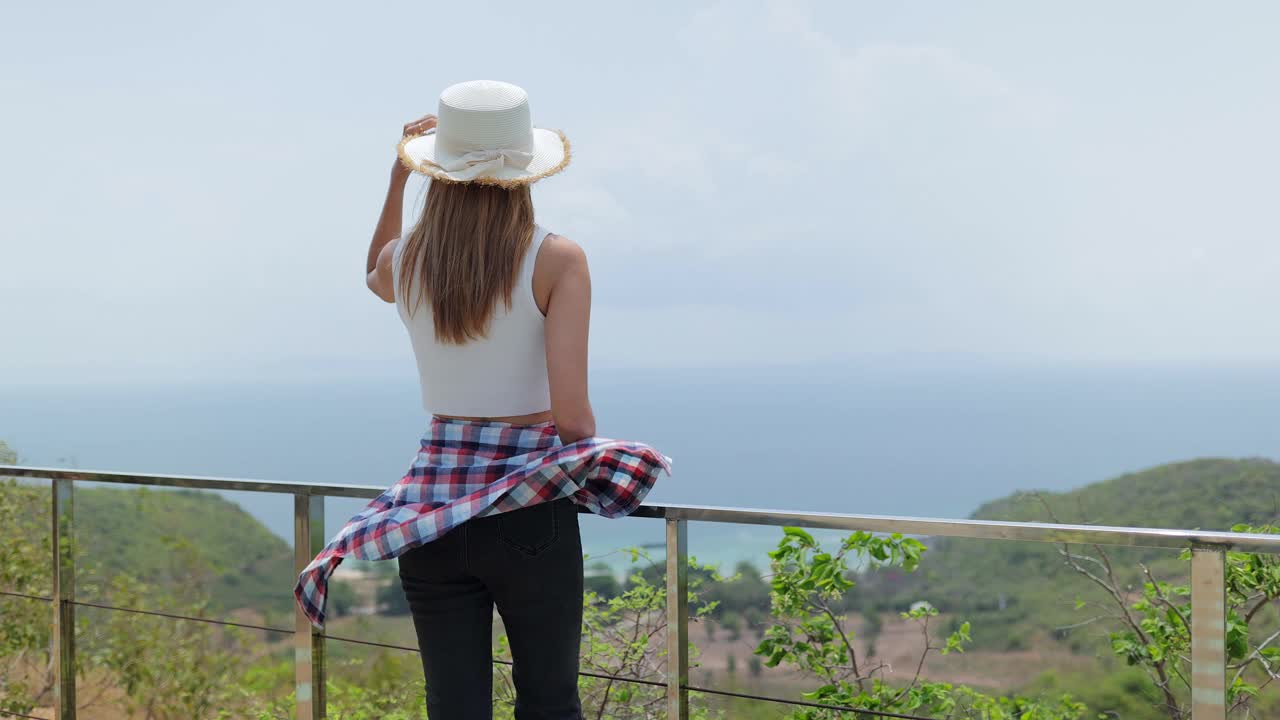 一位戴着帽子的女游客高高站在山上俯瞰大海。视频下载