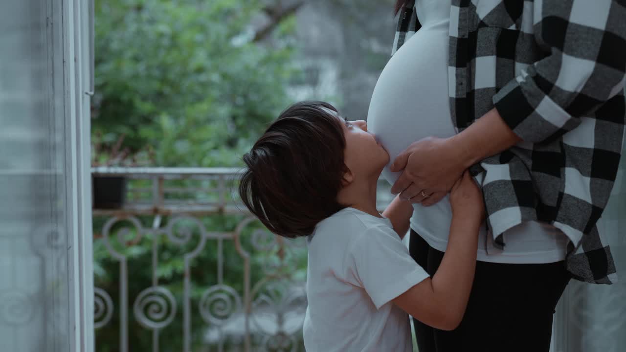 孕晚期，小男孩和妈妈站在阳台旁，亲吻着妈妈怀孕的肚子，表达对新生婴儿的爱视频素材