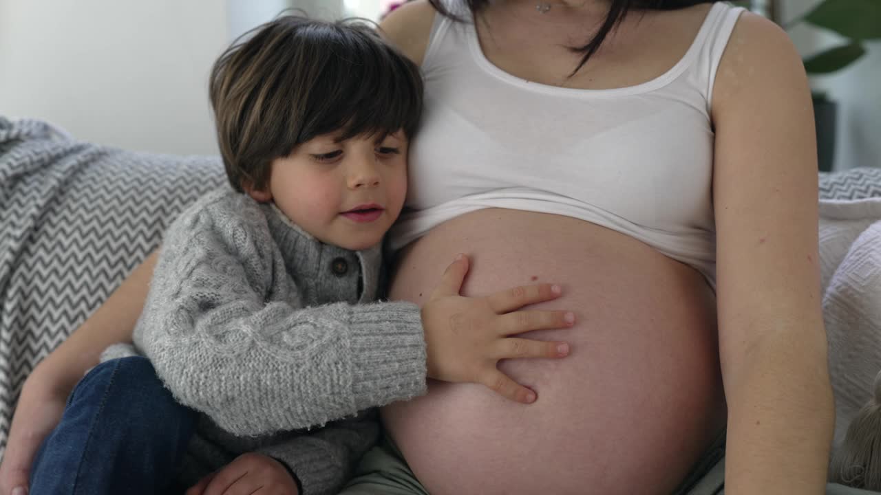 5岁男孩在怀孕后期温柔亲吻妈妈肚子的温馨时刻，孩子等待未出生的弟弟和妈妈坐在家里的沙发上，母爱的概念视频素材