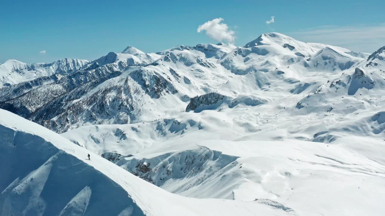 在晴朗的蓝天下，一个孤独的滑雪者从广阔的、白雪覆盖的山脉中一个原始的斜坡上滑下来视频素材