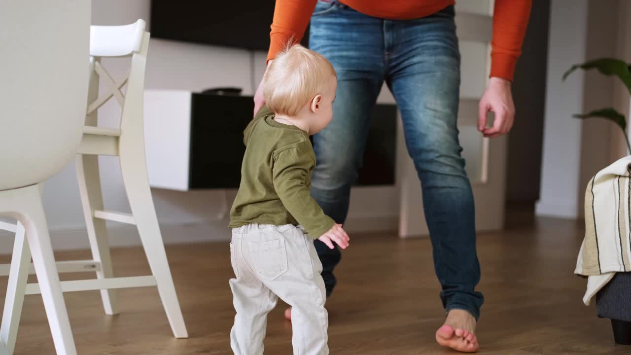 男人教婴儿走路。一岁的婴儿在父亲的支持下在室内迈出第一步。婴儿幸福，微笑着，看着镜头视频素材