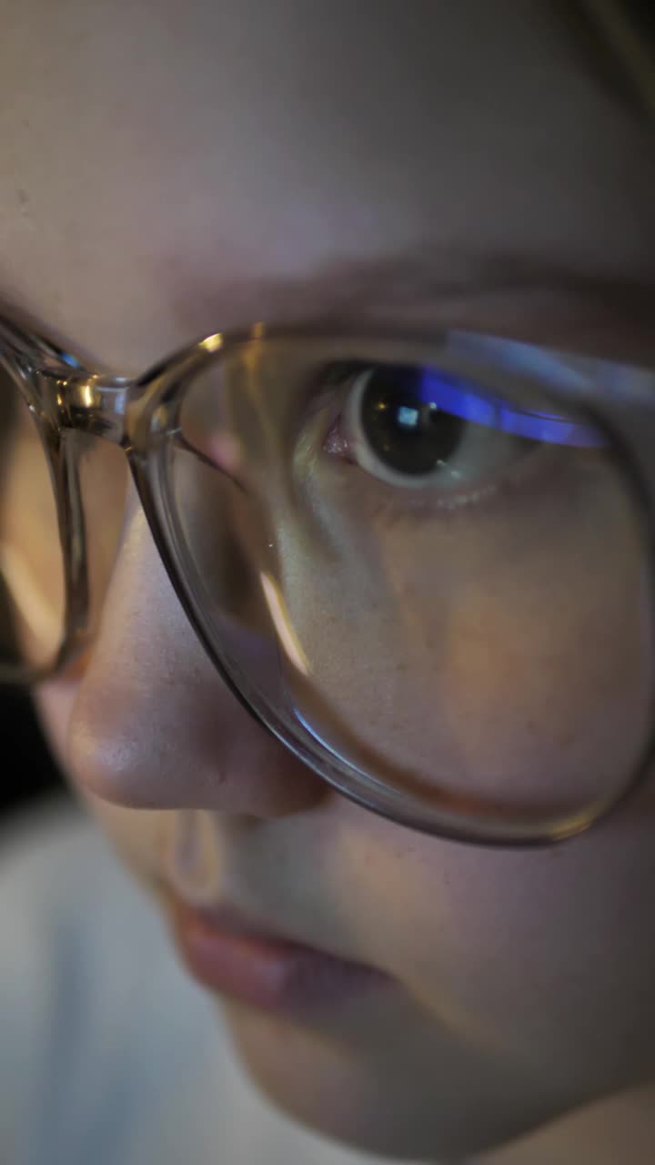 竖屏:8岁的小女孩戴着大眼镜，晚上看着电脑屏幕玩网络游戏。可爱的女孩在线教育。儿童网瘾视频素材