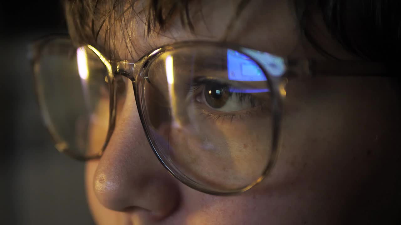 一个戴眼镜的聪明的11岁男孩在晚上盯着电脑显示器，一个戴眼镜的孩子在晚上玩网络游戏的特写。孩子们的电脑瘾。网络教育视频素材