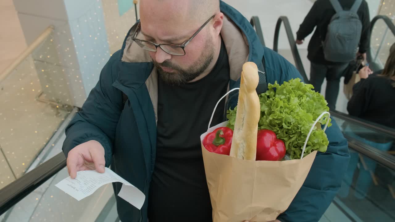 一个带着装满食物的购物纸袋的超重男子惊讶地看着超市自动扶梯上的纸质收据。男性对食品通胀和物价上涨感到震惊视频素材