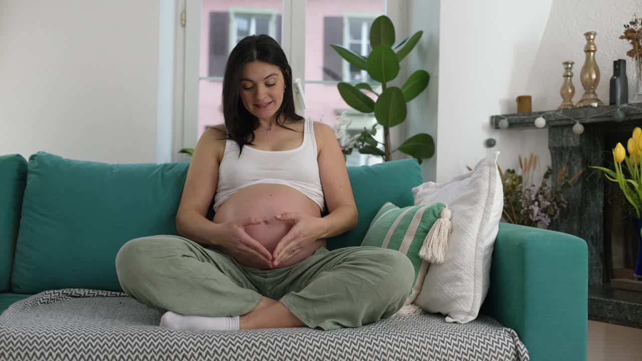 幸福怀孕的30多岁妇女坐在家里的沙发上，双手放在肚子上做心形符号，这是怀孕后期的肖像视频素材