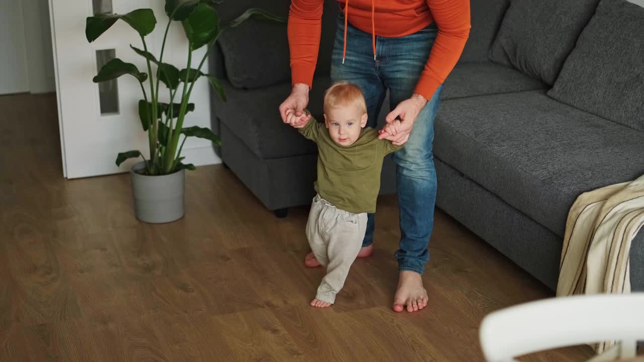 男人教婴儿走路。一岁的孩子在父亲的支持下在室内迈出第一步。婴儿幸福，微笑着，看着镜头视频素材